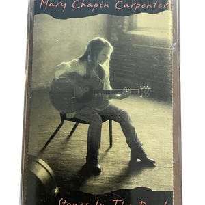 May include: A cassette tape case featuring a photo of Mary Chapin Carpenter playing a guitar. The text on the case reads "Stones In The Road".