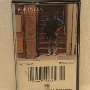 May include: A cassette tape with a black case and a white label. The label has a photo of Bob Dylan standing on a set of stairs. The text "BOB DYLAN STREET LEGAL" is printed in black on the label. The barcode number "74643 54534" is printed on the label. The text "COLUMBIA STEREO CASSETTE" is printed on the label.