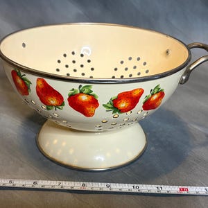 May include: Cream-colored metal colander with a decorative strawberry pattern. The colander has a silver rim, a handle, and a pedestal base. The bowl is perforated with small holes for draining. A measuring tape is in the foreground.