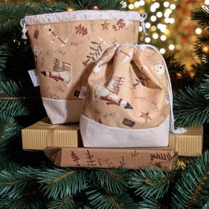 May include: Two beige fabric drawstring bags with a festive Christmas pattern of snowmen, trees, and stars. The bags have a cream-coloured base and are placed on wrapped gifts. The background is a Christmas tree.