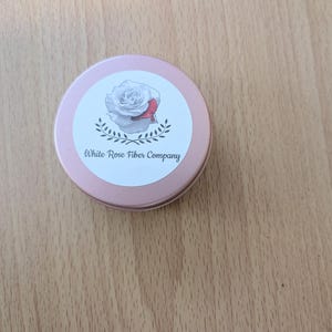 May include: A circular, pale pink tin with a white label displaying a watercolour rose and the text "White Rose Fiber Company." The tin rests on a wooden surface.