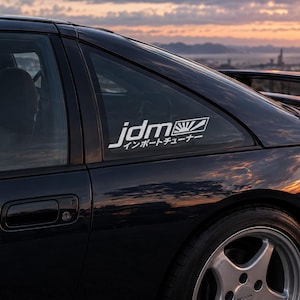 Könnte beinhalten: Ein dunkel lackierter Sportwagen mit einem weißen Vinyl-Aufkleber auf dem hinteren Seitenfenster. Der Aufkleber zeigt den Text jdm mit einer aufgehenden Sonne und japanischen Schriftzeichen darunter. Das Auto hat einen Heckspoiler und Alufelgen.