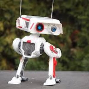 Könnte beinhalten: Ein weißes und rotes Spielzeugroboter mit blauen Augen und Antennen. Der Roboter hat vier Beine und steht auf einer grauen Oberfläche.