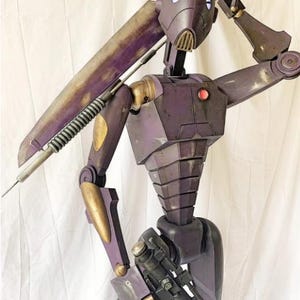 Könnte beinhalten: Eine lila und goldene Roboter-Actionfigur mit einer großen Waffe und einer kleineren Waffe. Der Roboter hat einen lila Helm mit einem roten Auge und einem schwarzen Visier.