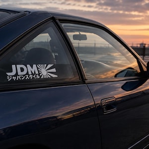 Könnte beinhalten: Ein dunkelblaues Auto zeigt einen weißen Vinyl-Aufkleber auf dem hinteren Seitenfenster. Der Aufkleber zeigt den Text JDM in fetten Buchstaben, eine stilisierte Sonnenstrahl-Grafik und die japanischen Schriftzeichen ジャパンスタイル.