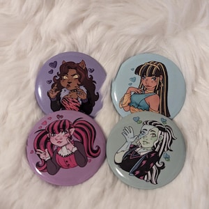 Puede incluir: Cuatro botones redondos con imágenes de personajes de dibujos animados. Los botones son morados, azules y azul claro. Los personajes son un hombre lobo, un ghoul, un zombie y un vampiro. Cada personaje tiene un diseño en forma de corazón a su alrededor.