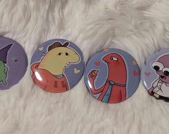 May include: Four round button pins with cartoon characters on a purple background. The characters are a green creature wearing a purple hat, a yellow creature wearing a red hoodie, a red creature with a blue tie, and a purple creature with large eyes wearing a white shirt.