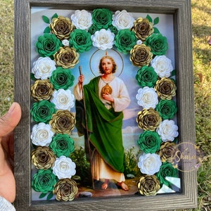 May include: A framed picture with a religious image of a man in a green robe holding a staff. The image is surrounded by a wreath of paper flowers in shades of green, white, and gold.