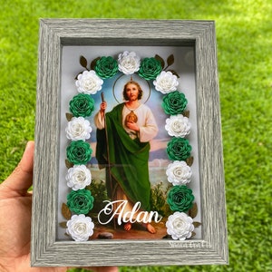 May include: A framed religious image of a saint, surrounded by white and green paper flowers. The image has a light gray frame and the name "Adan" is written in cursive. The background is a green lawn.