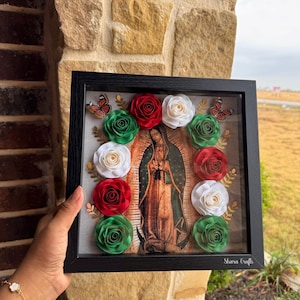 May include: A framed artwork featuring an image of Our Lady of Guadalupe, surrounded by red, white, and green paper roses. The black frame includes butterflies and the text "Sharia Crafts".