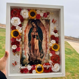 May include: A framed religious artwork featuring an image of the Virgin Mary, surrounded by red, white, and yellow paper flowers, including roses and sunflowers. The frame is white, and the artwork includes the text "Bendice este hogar".