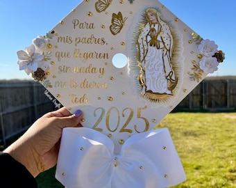 May include: White graduation cap decorated with gold glitter, white flowers, and a gold image of the Virgin Mary. The cap has a white bow with gold accents and the text "Para mis padres, que llegaron sin nada y me lo dieron todo 2025".
