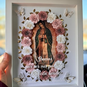 May include: A white frame with a picture of the Virgin Mary surrounded by paper flowers in shades of pink and white. The text "Gracias por ser mis padrinos Virgencita bendice y cuida a mis abuelitos" is written below the image.