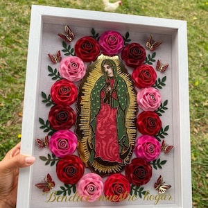 May include: A framed artwork featuring a religious image of the Virgin Mary surrounded by paper flowers in shades of pink and red. The text "Bendice nuestro hogar" is written in gold lettering at the bottom of the frame.