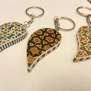 Puede incluir: Tres llaveros de madera con intrincados patrones geométricos en tonos marrones, dorados y azules. Cada llavero presenta un diseño en forma de lágrima con un anillo de llave plateado.