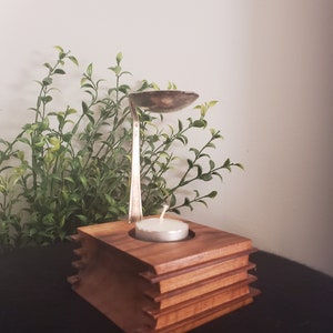 May include: A wooden candle holder with a stacked design and a silver spoon used as a diffuser. A white tea light candle sits in the holder.