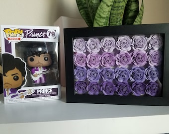 May include: A purple Pop Rocks vinyl figure of Prince holding a guitar, next to a framed picture of purple paper flowers.