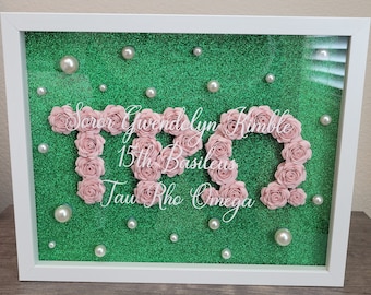 Può includere: Una scatola a ombre incorniciata con uno sfondo di glitter verde e fiori di carta rosa che compongono la scritta "T.R.O." con il testo "Soror Gwendolyn Kimble 15th Basileus Tau Rho Omega" in lettere bianche.