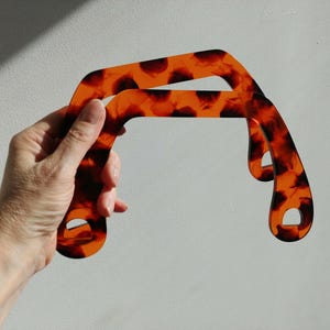 May include: Two brown and orange tortoise shell plastic handbag handles. The handles are shaped like a rounded arch.