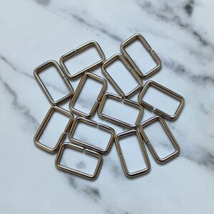 May include: A pile of silver-toned metal rectangular rings, also known as D-rings, used for attaching straps, chains, or other hardware to bags, purses, or other accessories.