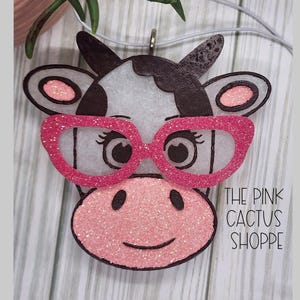 May include: A white and black cow with pink glitter glasses. The cow has a pink glitter nose and a pink glitter smile. The text "THE PINK CACTUS SHOPPE" is in the bottom right corner of the image.