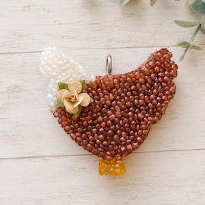 May include: A chicken-shaped pendant covered in brown and white beads with a small yellow beaded tail. A beige fabric flower is attached to the side of the chicken. A silver metal jump ring is attached to the top for hanging or attachment to a chain.