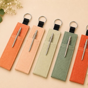 May include: Five rectangular fabric pen sleeves in shades of orange, peach, light green, sage green, and terracotta. Each sleeve features a silver metal pen tucked into a front pocket and a black fabric loop attached to a silver metal keyring.