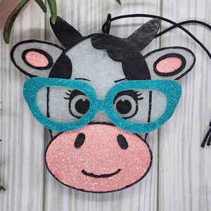 May include: A cow-shaped hanging ornament featuring a white face with black patches, a pink glittery snout, and bright turquoise glittery glasses. The ornament has black outlines, pink inner ears, and a black string attached for hanging purposes.