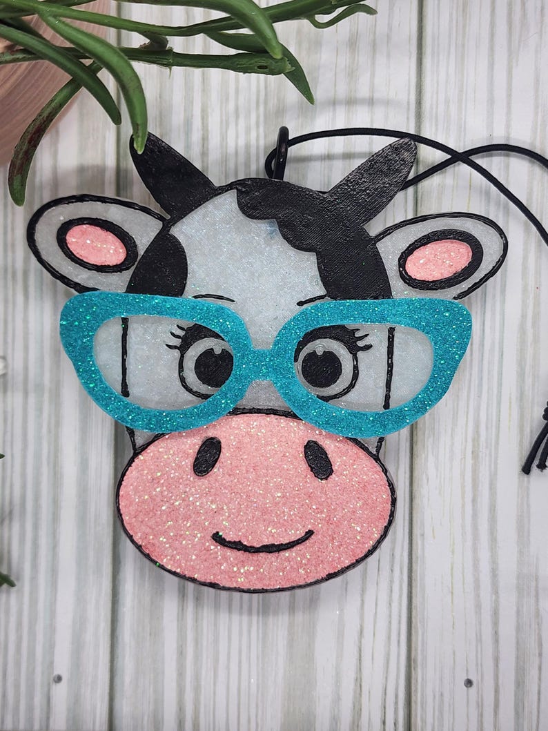 May include: A cow-shaped hanging ornament featuring a white face with black patches, a pink glittery snout, and bright turquoise glittery glasses. The ornament has black outlines, pink inner ears, and a black string attached for hanging purposes.