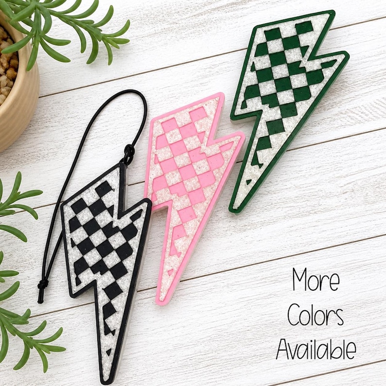 May include: Three lightning bolt-shaped accessories in black and white, pink, and green. Each has a checkered pattern and a white border. One black and white bolt is on a black cord. The text "More Colors Available" is also visible.