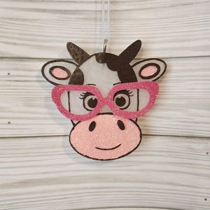 May include: A hanging ornament shaped like a cow head with a white face, black patches, and a pink snout. The cow features large, glittery pink glasses, black outlines, and pink inner ears, suspended by a thin white string for decorative display.