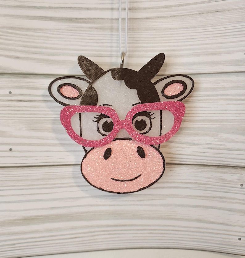 May include: A hanging ornament shaped like a cow head with a white face, black patches, and a pink snout. The cow features large, glittery pink glasses, black outlines, and pink inner ears, suspended by a thin white string for decorative display.