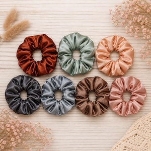May include: An assortment of eight satin scrunchies in various colors, including rust, sage green, peach, charcoal, silver, brown, and rose. The scrunchies are arranged on a light wood surface, with dried flowers and a crocheted item in the background.