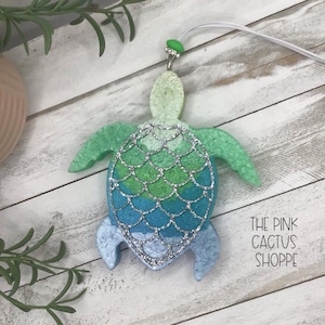May include: A sea turtle ornament in shades of green and blue with silver glitter accents. The ornament has a white string for hanging and a green gem at the top. The text "THE PINK CACTUS SHOPPE" is visible on the right side.
