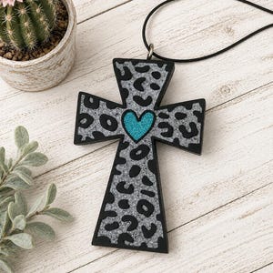 May include: A cross-shaped pendant featuring a silver glitter background with a black leopard print pattern and a central teal glitter heart. The pendant is attached to a thin black cord, creating a necklace that displays a bold contrast.