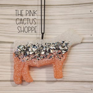 May include: A white and pink cow silhouette ornament with silver glitter and a black string for hanging. The ornament has the text "THE PINK CACTUS SHOPPE" on the top.
