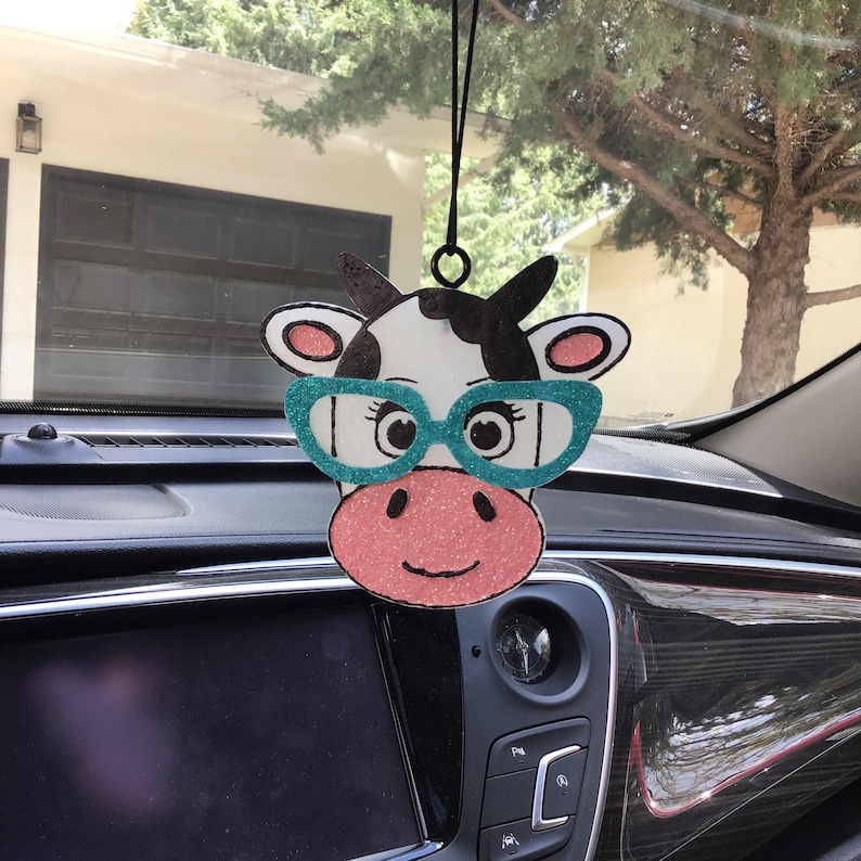 May include: A hanging car air freshener shaped like a cow head with black and white markings. The cow features glittery pink ears and a glittery pink snout, along with bright teal glittery glasses. It hangs from a black string loop.