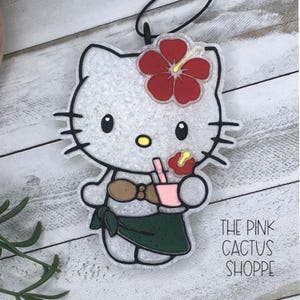 May include: A white cat-shaped air freshener with a red hibiscus flower and a pink drink. The cat wears a brown top and a green skirt. The text "THE PINK CACTUS SHOPPE" is printed on the bottom right.