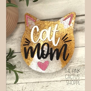 May include: A cat-shaped sign with the words "Cat Mom" in white and black lettering. The sign has a gold and silver textured background, pink accents, and a pink heart. The sign also includes the text "THE PINK CACTUS SHOPPE".