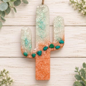 May include: A cactus-shaped wall hanging made of resin with a gradient color transition from a translucent white and teal glitter top to a solid orange base, accented with small, smooth, embedded turquoise-colored stones along the center.