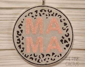 May include: A circular hanging ornament with a white glittery base featuring a black leopard print pattern. The word MAMA is centered in large, rose gold glitter letters. A black cord is attached to the top for hanging purposes.