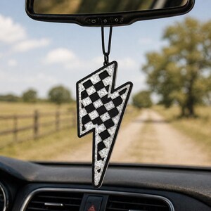 May include: A black and white checkered lightning bolt car air freshener hanging from a rearview mirror. The air freshener has a black border and a black string for hanging. The background shows a dirt road and a field.