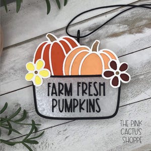 May include: A decorative hanging ornament featuring two orange pumpkins in a white container with the text FARM FRESH PUMPKINS in black lettering. The design includes a yellow flower on the left and a dark brown flower on the right, suspended by a black cord.