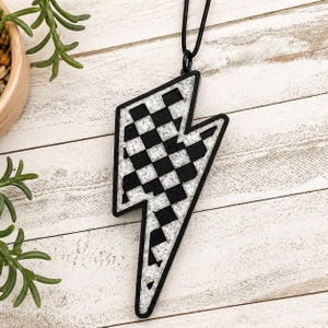 May include: A black and white checkered lightning bolt pendant necklace. The pendant has a black outline and a silver glitter-like fill. The necklace cord is black.