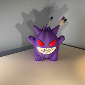 May include: A purple 3D printed Ghost Pokemon character desk organizer with red eyes and a toothy grin. The organizer is holding several pens and paper clips.