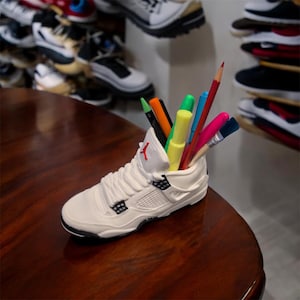 May include: A white sneaker-shaped desk organizer with a red logo, filled with colorful pens, pencils, and a paintbrush. The organizer sits on a dark wooden surface, with a blurred background of shoes.