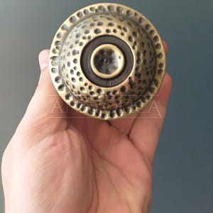 May include: A round, brass-colored door knocker with a hammered texture and a circular, black center.