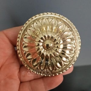 May include: A gold-coloured, circular decorative knob with a floral design. The knob has a central, raised button surrounded by petal-like shapes and a textured border. The word "ANATOLICA" is printed on the knob.