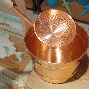 May include: A copper bucket with a hammered finish and a matching copper ladle with a wooden handle.