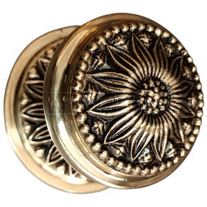 May include: Two antique brass door knobs with a sunflower design. The knobs are round with a detailed floral pattern and a beaded border. The brass has a warm, golden hue, and the design is raised, creating a textured surface.